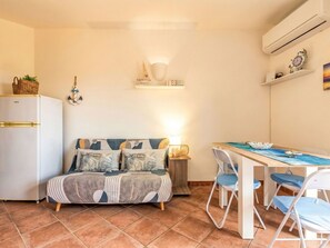 A compact living space with a kitchenette, a sofa with patterned cushions, a dining table with chairs, and a wall-mounted air conditioner.