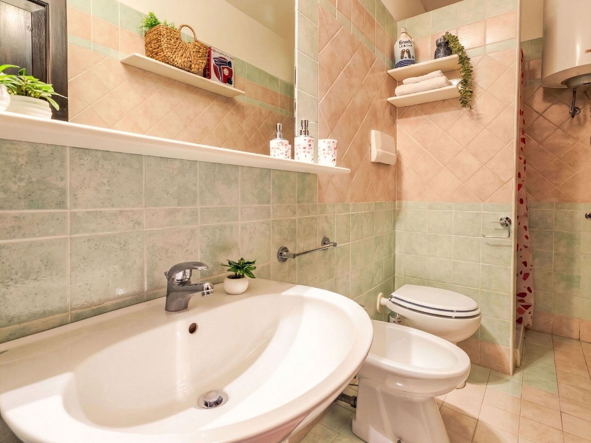 Une salle de bain équipée d’un lavabo, de toilettes et d’une douche. On y trouve des étagères avec des produits de toilette et une plante.