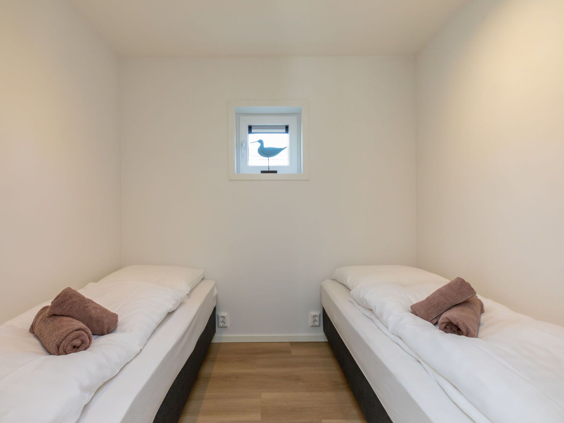 A small, clean room with two single beds, a window with a bird decoration, and a wooden floor.