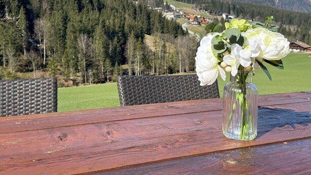 Una mesa de madera con un florero de vidrio transparente con flores blancas, frente a un fondo de montañas nevadas y un valle verde.