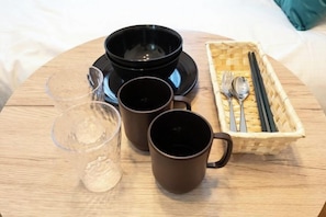 Une table ronde en bois, sur laquelle se trouvent un bol noir, deux tasses brunes, un verre contenant une boisson et un panier tressé rempli de couverts.