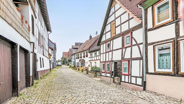 Une rue pavée bordée de maisons traditionnelles à colombages, un regard d’égout et une clôture métallique sur la gauche.