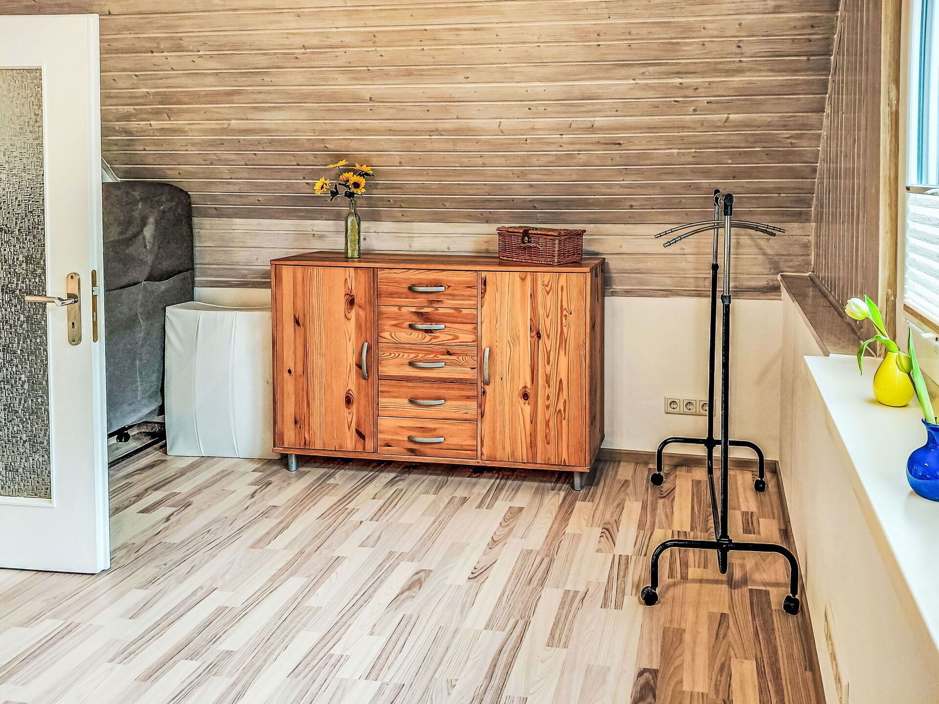 Un mueble de madera con cajones, un jarrón con flores y una lámpara de pie en una habitación con suelo de madera y una ventana.