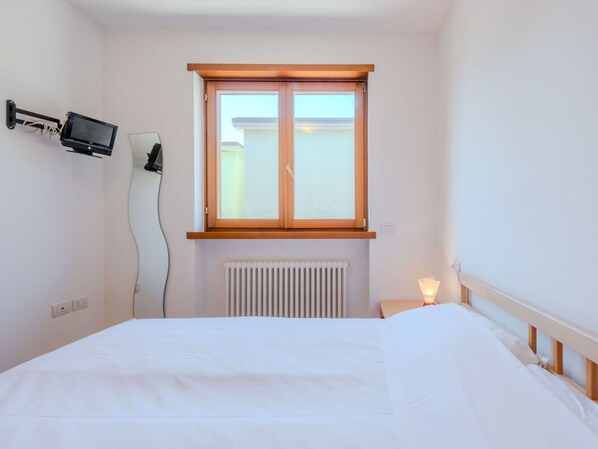 A bedroom with a bed, a window, a wall-mounted TV, a radiator, and a bedside lamp.