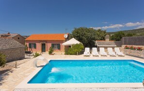 Une piscine entourée de chaises longues, un bâtiment en pierre avec un toit rouge et un ciel bleu azur.