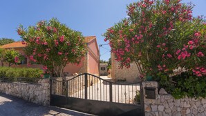 A gated entrance with stone walls and a metal gate, leading to a street with buildings and flowering trees.