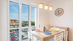 Un comedor con una mesa preparada para cuatro personas, vistas a coches aparcados a través de amplias ventanas, y un reloj en la pared.