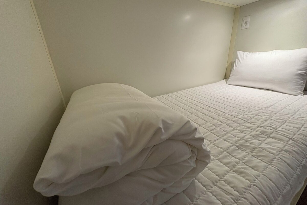 A neatly made bed with a white quilted bedspread and pillows, in a room with plain walls and a simple, functional design.
