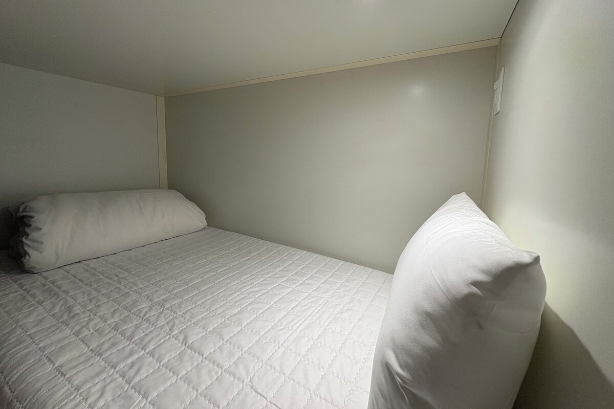 Ein kleines Zimmer mit einem Einzelbett, einer schlichten weißen Wand und Decke, wenigen Möbeln und einer Deckenleuchte.
