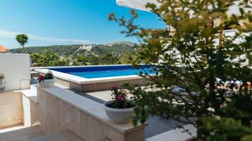A swimming pool with a view of a hilly landscape, surrounded by potted plants and a white umbrella.