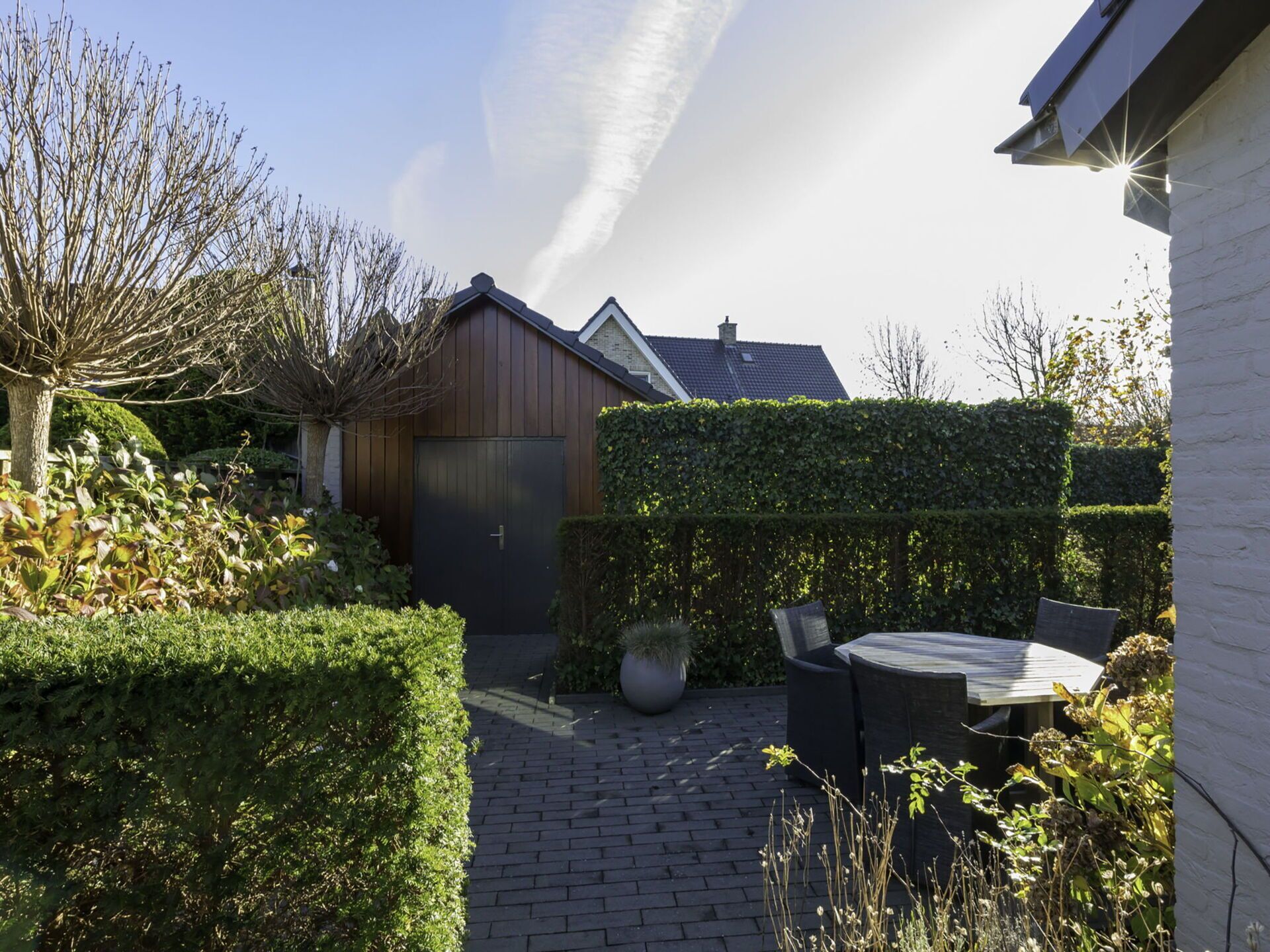 Een geplaveid tuinpad dat leidt naar een houten tuinhuis, omringd door netjes gesnoeide hagen en een bloempot.
