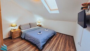 Une chambre avec un plafond en pente, un puits de lumière, un lit recouvert d’une couvre-lit à motifs bleus, deux lampes de chevet, un téléviseur fixé au mur et un plancher en bois.