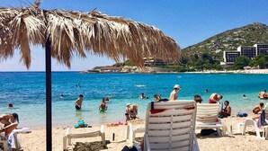 A beach with clear blue water, people swimming and sunbathing, and a mountain in the background.
