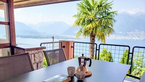 Una mesa con un libro, una cafetera y tazas en un balcón con vista a un lago y montañas.