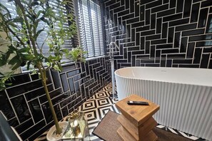 A modern bathroom with a black and white geometric tile backsplash, a white bathtub, and a wooden side table with a remote control. - Soho Heritage House Chinatown Bangkok (Bangkok)