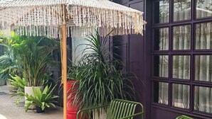 Un espace patio agrémenté d’un parasol en fibres tressées, de chaises vertes, de plantes en pot et d’une grande fenêtre offrant une vue dégagée.