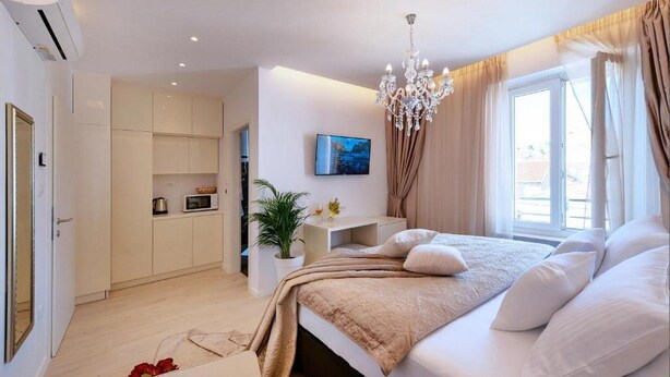 Un dormitorio moderno con una cama grande, una araña, un televisor y una kitchenette al fondo.