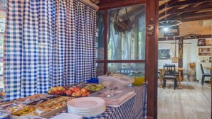 Un bufé de desayuno con diversos alimentos y utensilios, un mantel a cuadros y vista al comedor a través de un amplio ventanal.