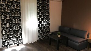 Habitación con sofá, una mesita y una planta en maceta. Se aprecia una ventana con cortinas de encaje y un cortinaje estampado. Un equipo de aire acondicionado está fijado a la pared.