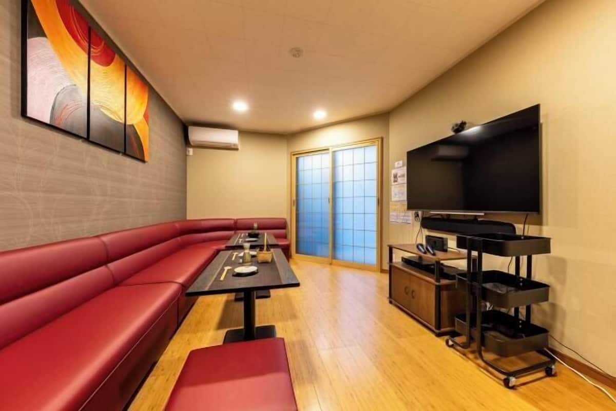 Una habitación con un sofá rojo, una mesa con un control remoto, un televisor sobre un mueble bajo y una puerta corredera.