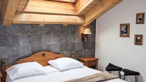 Une chambre avec un plafond en pente, un lit recouvert de draps blancs, une tête de lit en bois, une table de chevet, une lampe et des tableaux encadrés accrochés au mur.
