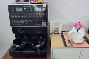 Une machine à café avec deux verseuses, un plateau avec des tasses et un parapluie rose, ainsi qu’une boîte de café instantané, le tout posé sur une table.