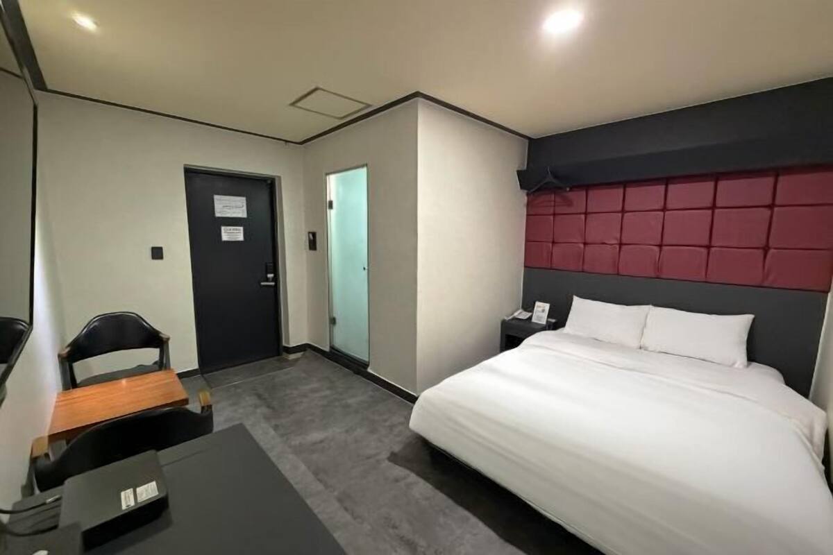Quarto de hotel com cama, escrivaninha, cadeira, mesa de cabeceira e porta.