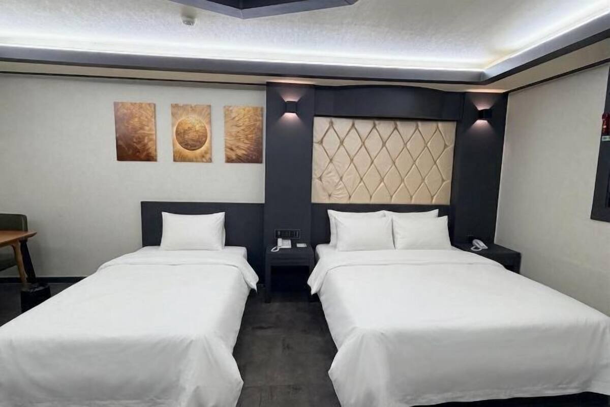 Quarto de hotel com duas camas, cabeceira com padrão de losangos, uma mesa pequena e obras de arte na parede.