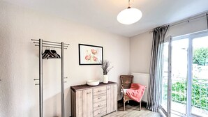 Une chambre moderne avec un plancher en bois, un porte-manteau, une commode, une chaise avec un coussin rose, une plante en pot et une fenêtre donnant sur un paysage verdoyant.