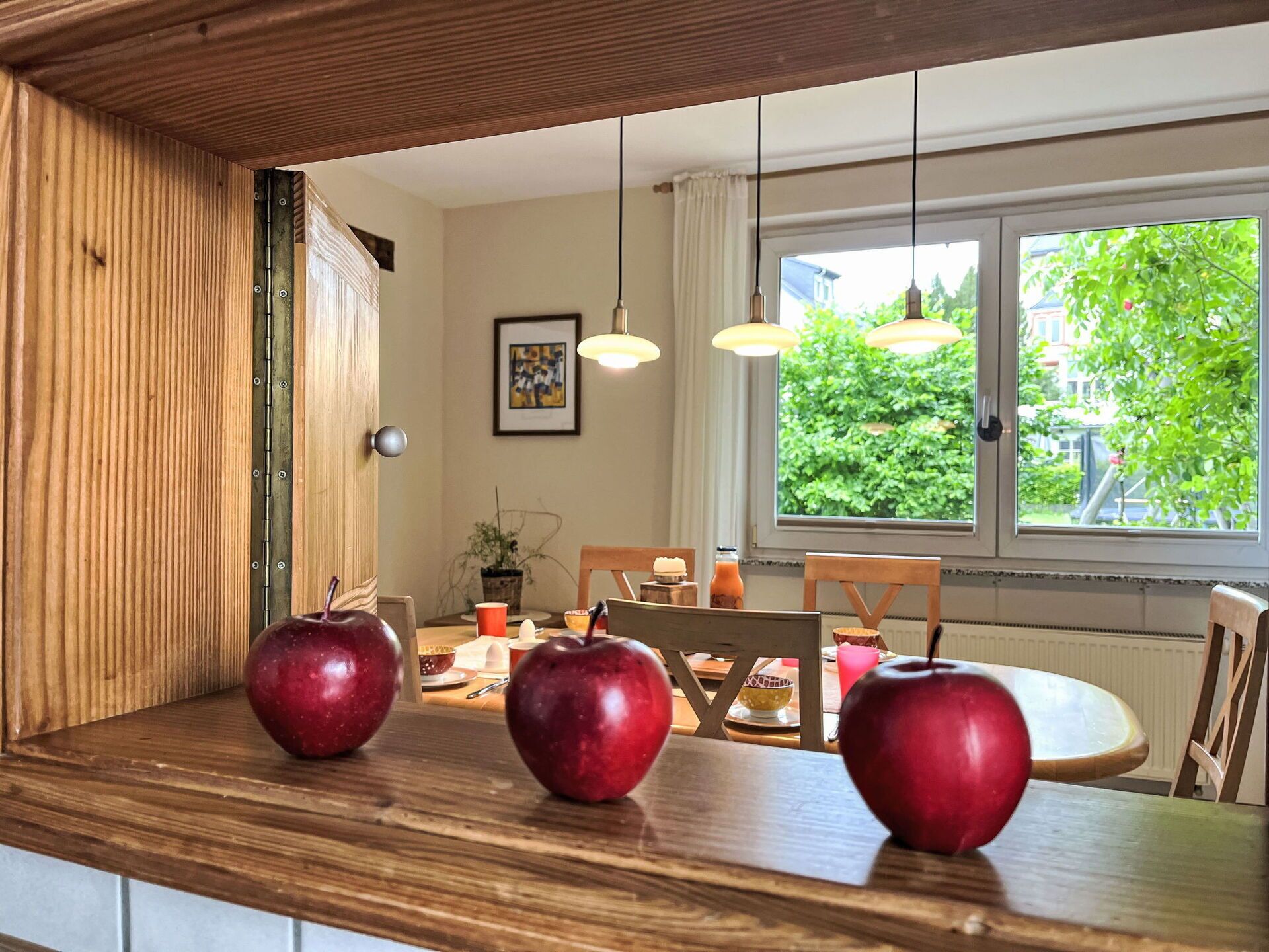 A wooden dining table with three apples, a framed picture on the wall, and a window with a view of greenery.