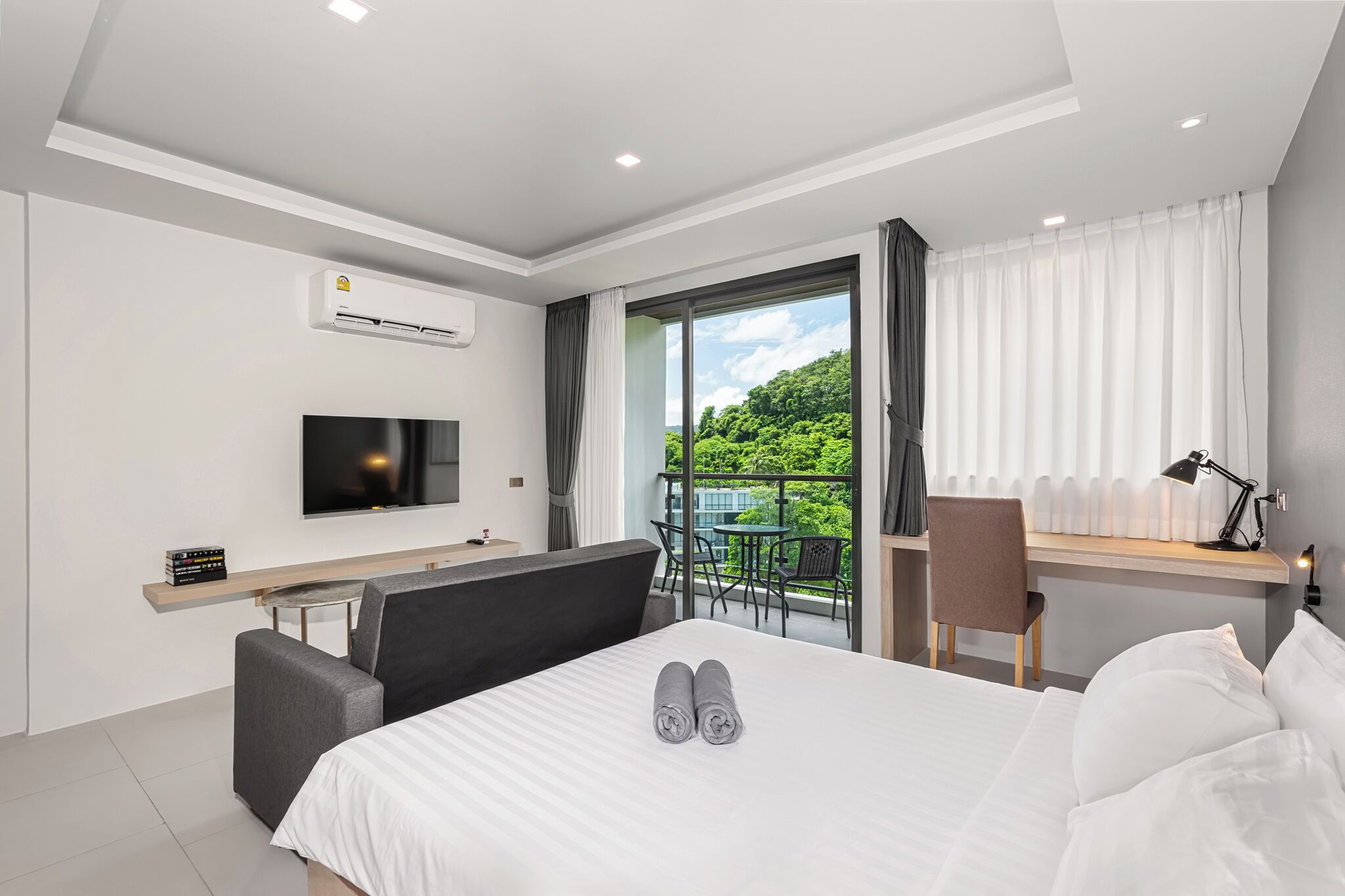 Une chambre d’hôtel moderne équipée d’un lit, d’un bureau, d’une télévision et d’un balcon offrant une vue sur la verdure.
