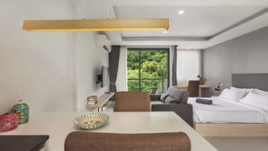 A modern hotel room with a large bed, a desk with a chair, a dining table, and a view of greenery outside.