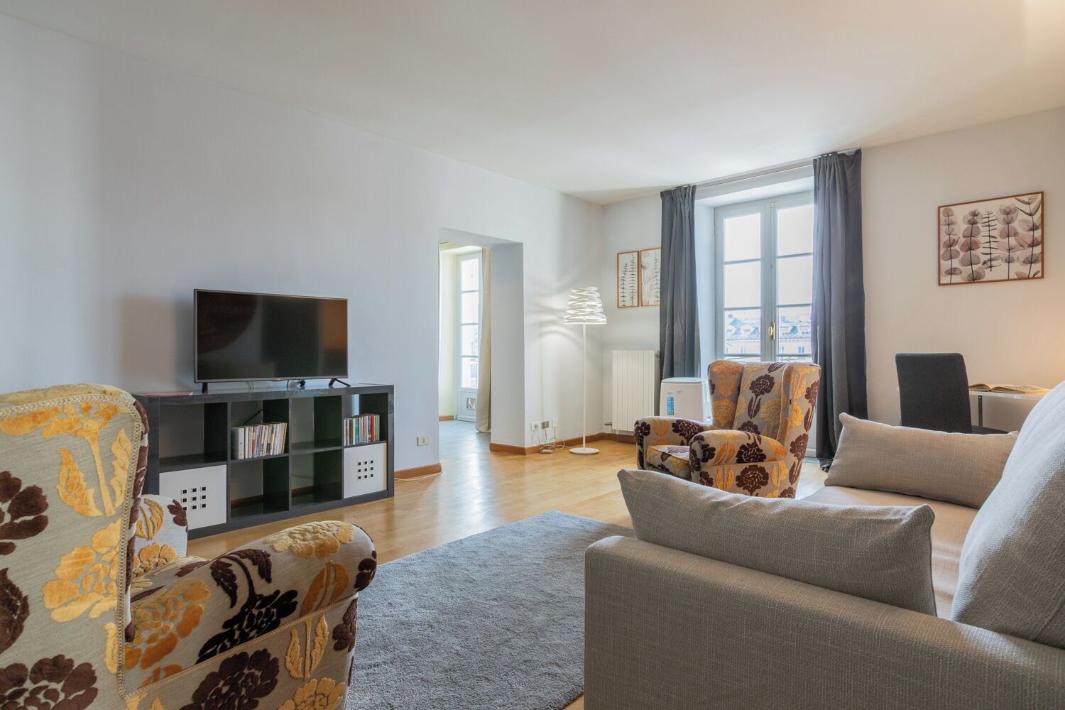 Una sala de estar con televisor de pantalla plana, estantería, sillones estampados, lámpara de pie y ventana con cortinas.