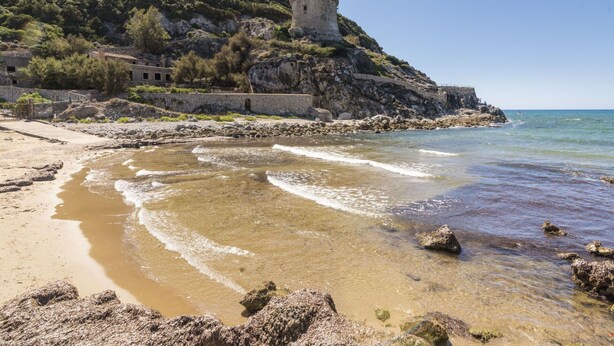 A coastal scene with a stone tower on a cliff, clear waters, and rocky beach.