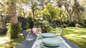 An outdoor dining table set with plates, glasses, and cutlery, surrounded by lush greenery and trees.