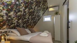Quarto com teto inclinado, cama com roupa de cama branca, mesa de cabeceira com abajur e um mural de parede com motivos florais.
