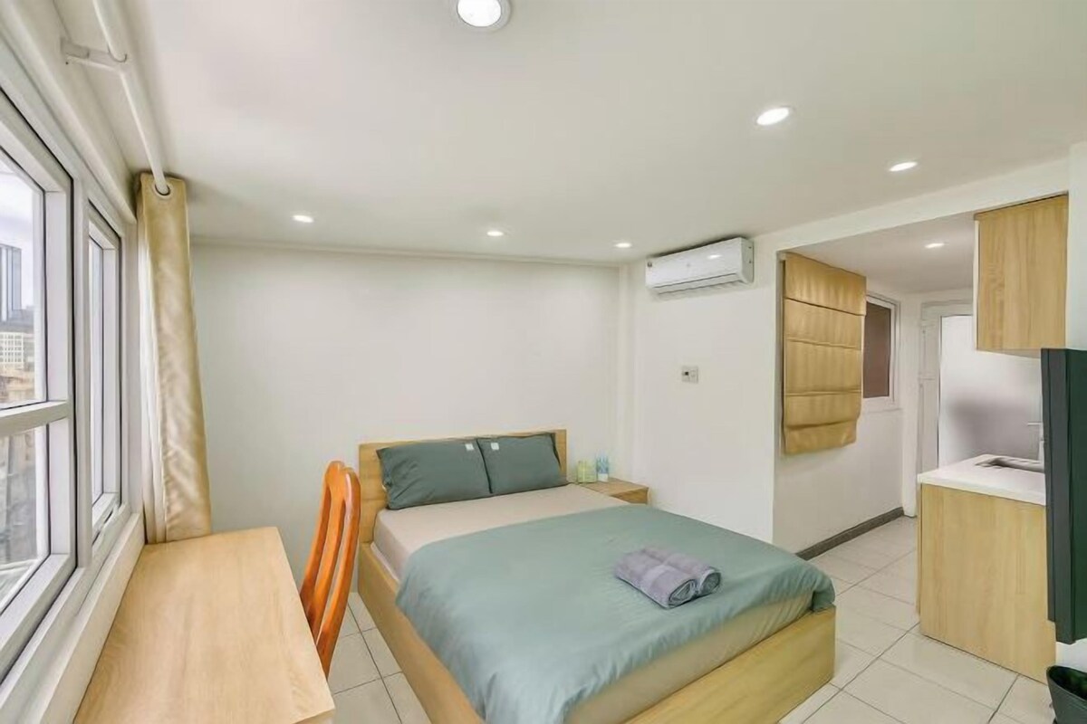A bedroom with a bed, a desk, a chair, a window, and a kitchenette.
