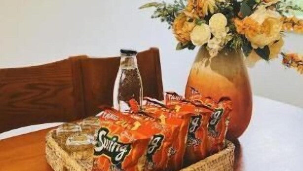 A dining table with a woven tray holding a pack of Snickers bars, a vase with flowers, and a bottle.