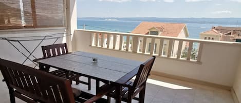 A balcony with a table and chairs, a view of the sea and buildings, and a window with blinds.