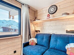 Ein gemütliches Zimmer mit einem blauen Sofa, einer Holzwand, einer Uhr und einem Fenster mit Blick auf ein Gebäude.