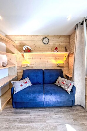 A room with a blue sofa, wooden shelves, a clock, and a window with curtains. - Les Olympiades by Interhome (Val Thorens)
