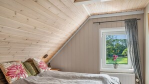 A bedroom with a sloped ceiling, a bed with a grey bedspread, two pillows, a nightstand, a window with a view of greenery, and a radiator.