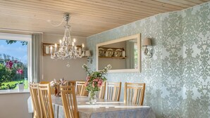 A dining room with a chandelier, wooden chairs, a table with a lace tablecloth, and a view of a garden through a window.