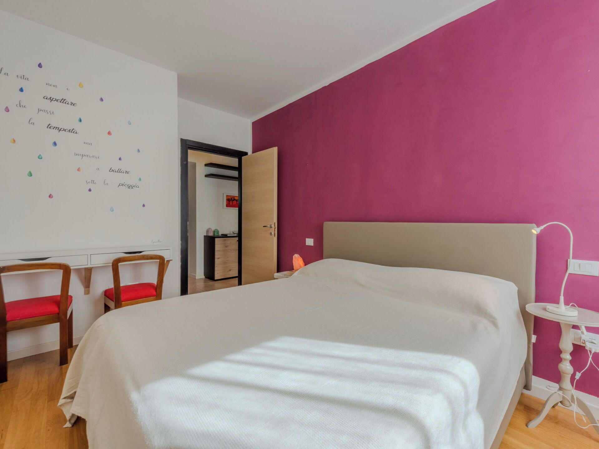 A bedroom with a bed, a desk, two chairs, a small table, and a wall with a decorative item.