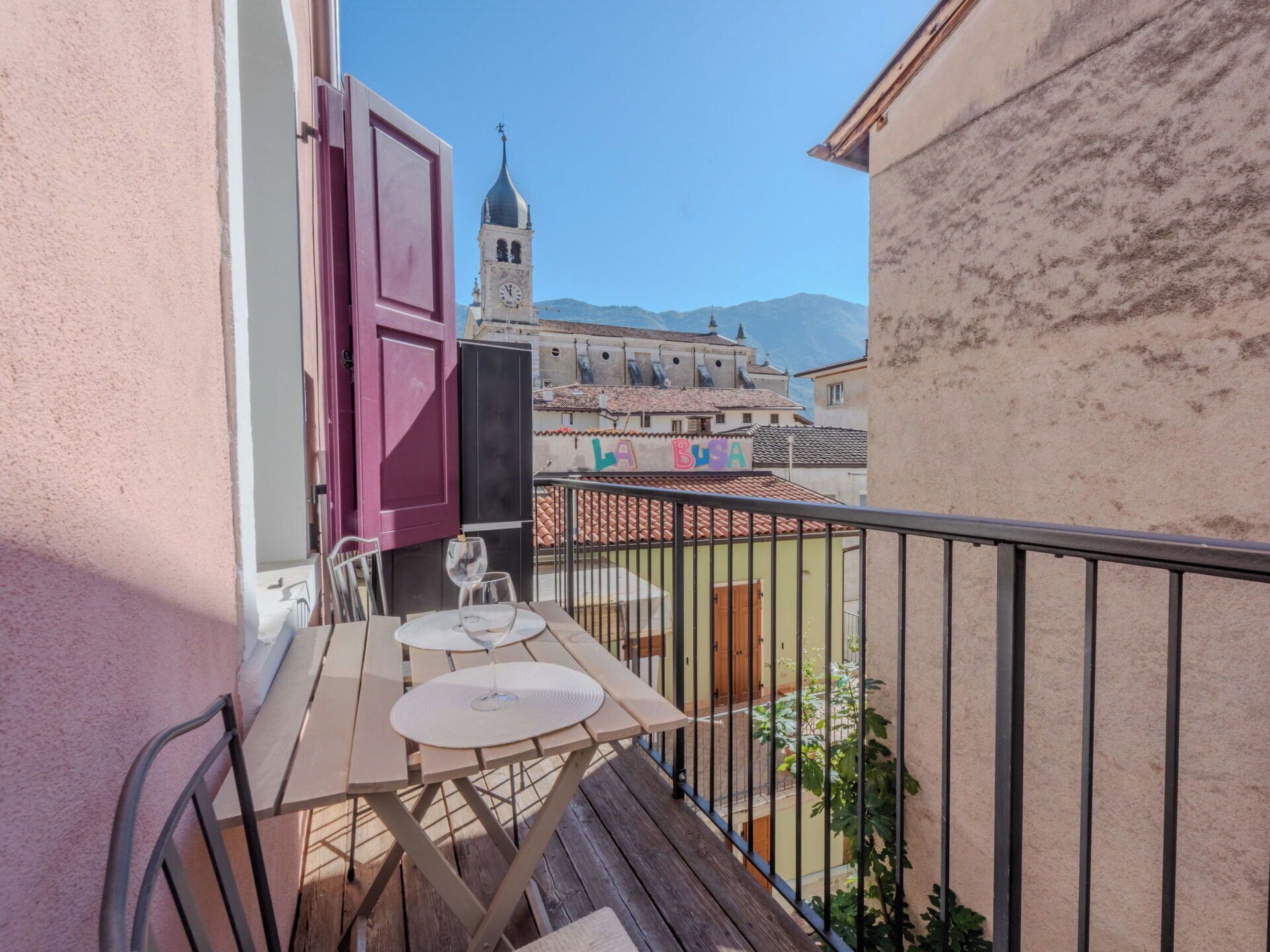 Un balcon avec une table et des chaises, un volet rouge et une vue sur un bâtiment historique doté d’une tour horloge.