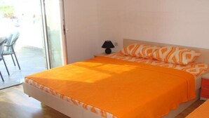 Ein Schlafzimmer mit einem Bett mit orangefarbener Bettwäsche, einem Nachttisch mit Lampe und einem Stuhl, der durch eine Glastür sichtbar ist.