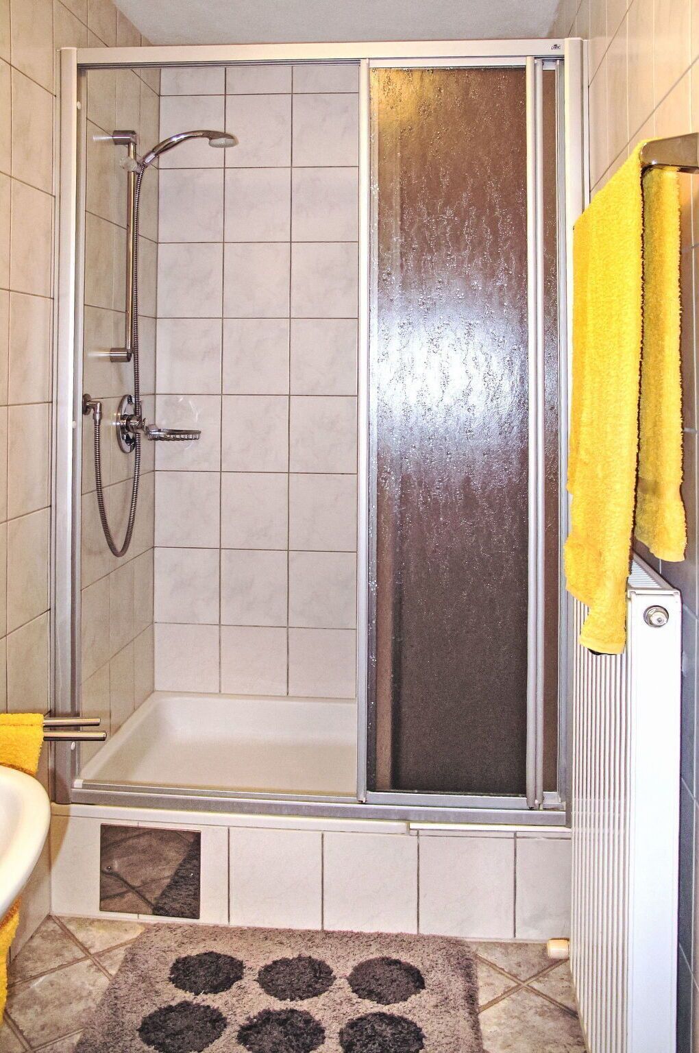 A shower enclosure with a glass door, a yellow towel on the radiator, and a patterned rug on the floor.