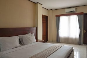 A hotel room with a bed, a wooden headboard, an air conditioning unit, a door, and a window with curtains. - Mahajaya Hotel Denpasar (Bali)