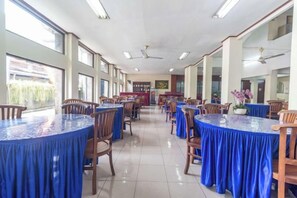 A spacious dining area with round tables covered in blue tablecloths, wooden chairs, and large windows allowing natural light. - Mahajaya Hotel Denpasar (Bali)