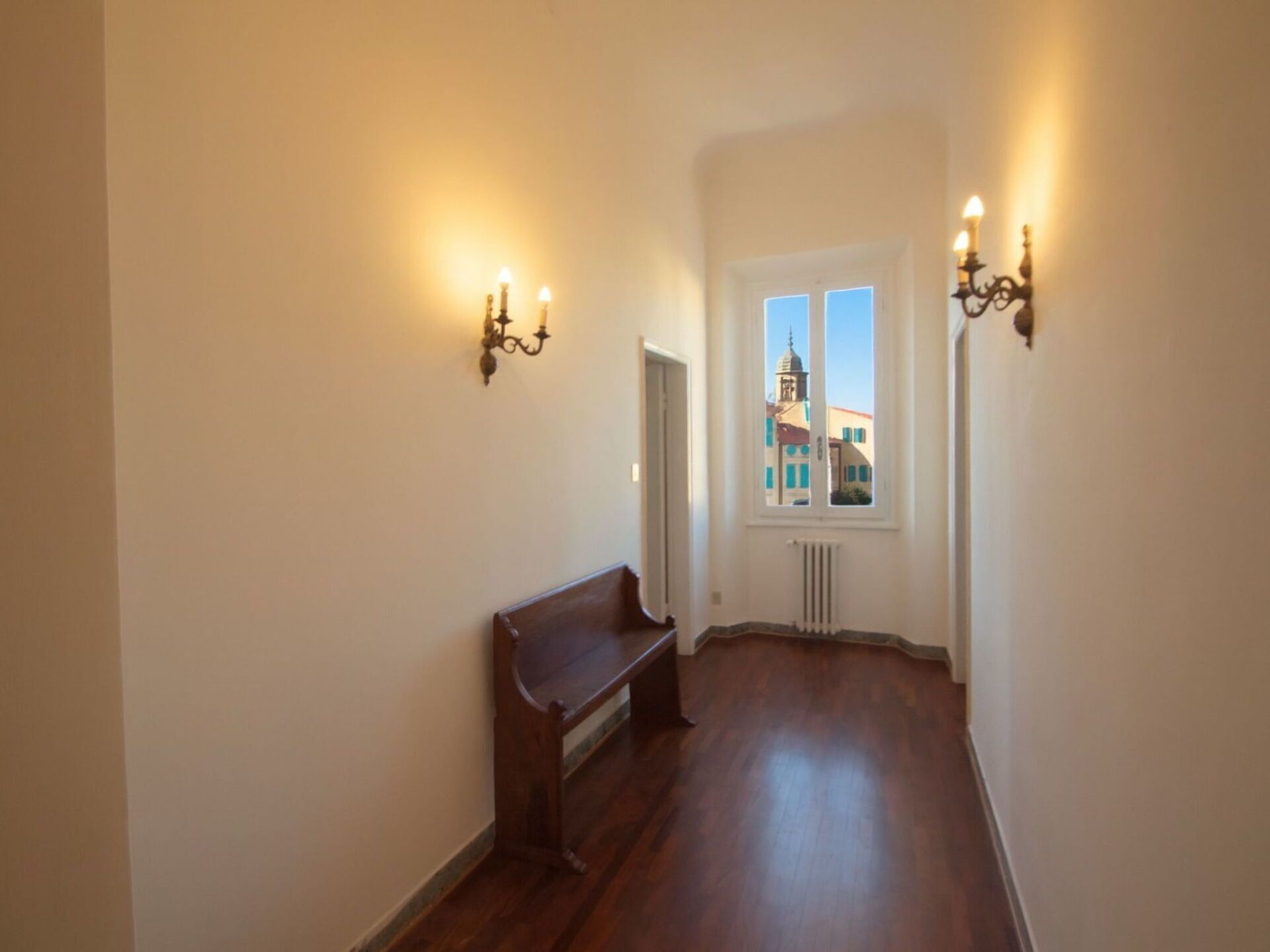 A narrow hallway with wooden flooring, a bench, and two wall-mounted sconces. A window with a view of buildings is visible in the background.